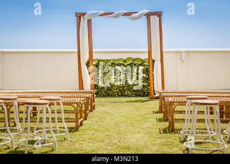 Nozze in legno arch per una cerimonia decorata con tessuto bianco, fiore parete con la parola amore fatta con fiori tropicali, sedie di rattan e panchine. Foto Stock