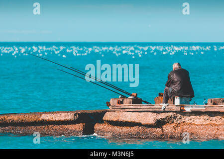 Un anziano uomo solitario è la pesca su un molo sul mare. La vecchiaia e la solitudine. Foto Stock