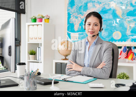 Professional bella donna d'affari di indossare le cuffie lavorando su agente di viaggi aziendali e mani incrociate di fronte alla telecamera sorridente. Foto Stock