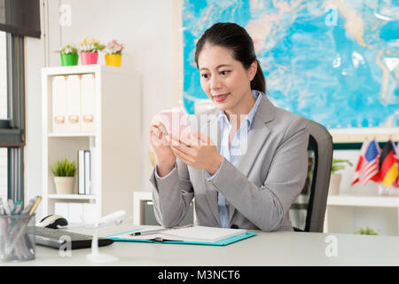 Bella bella femmina travel agent manager seduto nel lavoro di ufficio e utilizzando mobile smartphone verifica prenotazione online dal cliente. Foto Stock