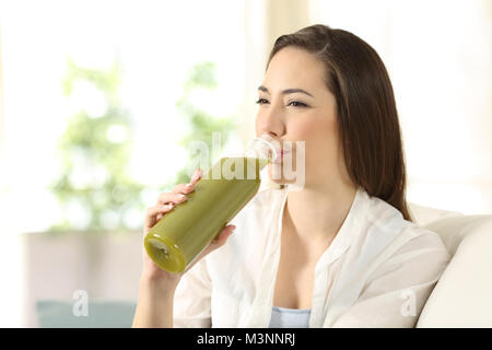 Donna felice di bere un succo vegetale da una bottiglia seduta su un divano nel salotto di casa Foto Stock