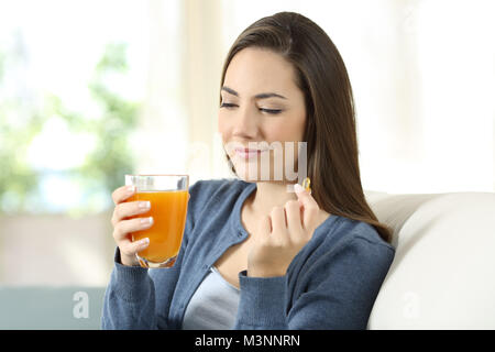 Donna che mantiene un succo di arancio e vitamina pillola seduta su un divano nel salotto di casa Foto Stock