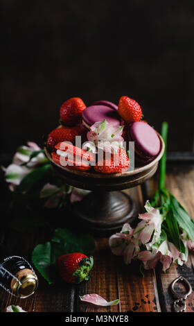 Ciotola con macarons e fragole su un tavolo di legno con fiori e bottiglia di champagne Foto Stock