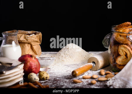 Vista ravvicinata di deliziosi biscotti fatti in casa in una bottiglia di vetro e gli ingredienti per produrre un impasto sul piano del tavolo su nero Foto Stock