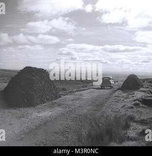 Degli anni Cinquanta, storico, automobile parcheggiata su un vicolo del paese betwee due grandi pile di torba, Irlanda del Nord. Foto Stock