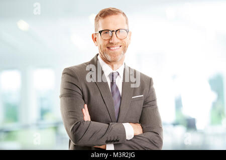 Senior Investment Advisor imprenditore con bracci ripiegati indossare tuta mentre in piedi presso l'ufficio e guardando la fotocamera. Foto Stock