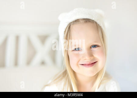 Ritratto carina ragazza bionda. Smilling capretto nel cappello bianco. Bambino sul letto in camera luminosa. Adorable baby indossando cappello spiritoso con piccole orecchie. Infanzia felice concetto. Soddisfatti i bambini Foto Stock