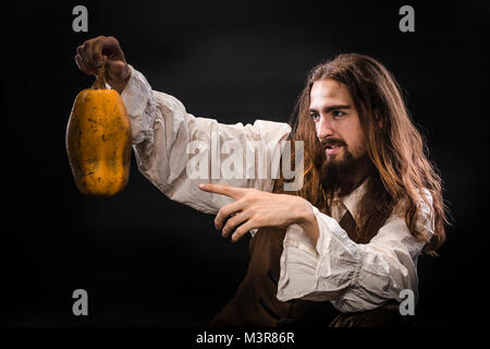 Ritratto di un uomo con la barba e capelli lunghi che indossa un medievale costume pirata su uno sfondo nero, un pirata tenendo una zucca matura