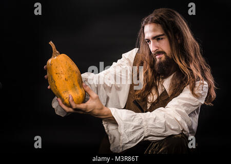 Ritratto di un uomo con la barba e capelli lunghi che indossa un medievale costume pirata su uno sfondo nero, un pirata tenendo una zucca matura