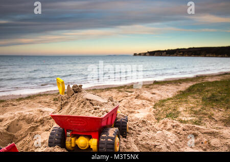 Close up di un bambino giocattolo i carrelli abbandonati nella sabbia, su una deserta spiaggia del Regno Unito. Di sera presto la luce del sole crea colori sorprendenti attraverso il cielo. Foto Stock