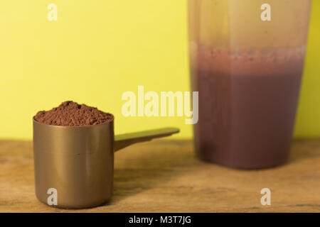 La proteina del siero di latte in polvere bodybuilding sportivi supplemento. Close up di scoop con il cioccolato in polvere e shaker su tavola di legno. Colore di sfondo: giallo. Foto Stock