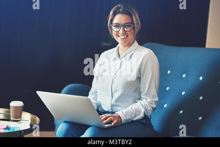 Femmina giovane imprenditore sorridente con fiducia mentre è seduto su un divano a lavorare da casa utilizzando un computer portatile Foto Stock