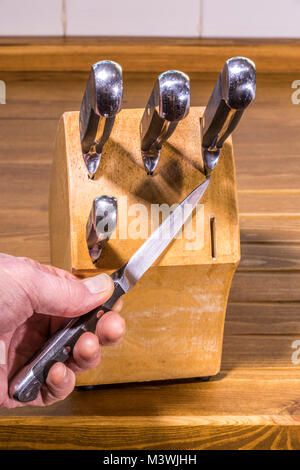 Mano d'uomo tenendo un affilato coltello in acciaio, uno dei cinque in un set. Le lame dei coltelli resto negli slot in un pesante blocco di legno su un piano di lavoro di pino. Regno Unito. Foto Stock