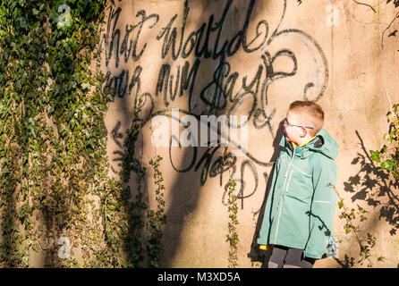 6 anno vecchio ragazzo con gli occhiali in piedi accanto a vernice spray i graffiti sulla parete, nel quale si legge che cosa sarebbe la vostra mamma dire?, Scotland, Regno Unito Foto Stock