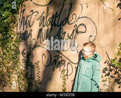 6 anno vecchio ragazzo con gli occhiali in piedi accanto a vernice spray i graffiti sulla parete, nel quale si legge che cosa sarebbe la vostra mamma dire?, Scotland, Regno Unito Foto Stock