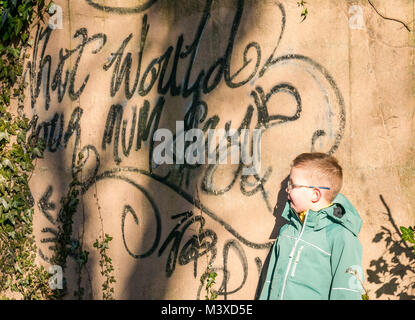 6 anno vecchio ragazzo con gli occhiali in piedi accanto a vernice spray i graffiti sulla parete, nel quale si legge che cosa sarebbe la vostra mamma dire?, Scotland, Regno Unito Foto Stock