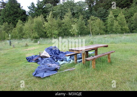 Abandonned, crollato, blu tenda, sacco a pelo e camping sedile accanto al legno un tavolo da picnic sul prato verde. Camping disater. Foto Stock