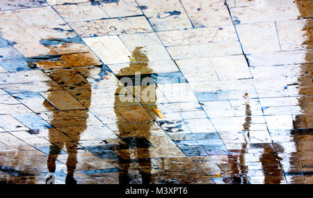 Sfocata Rainy day, le persone camminare sotto ombrellone silhouettes di riflessione sul bagnato city square in alto contrasto Foto Stock