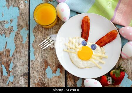 Colazione di pasqua con cute bunny faccia fatta di uova e bacon. Tabella scena, sopra vista su un rustico in legno blu sullo sfondo. Foto Stock