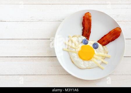 Colazione di pasqua con cute bunny faccia fatta di uova e bacon. Sulla piastra bianca contro un bianco sullo sfondo di legno. Foto Stock