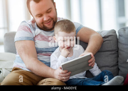 Immagine di papà con suo figlio con la compressa seduto sul divano grigio Foto Stock