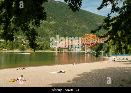 Nelson, British Columbia, Canada: rotante parco sul lago spiaggia pubblica su un pomeriggio d'estate, autostrada 3A ponte al di sopra del braccio ovest del Lago Kootenay. Foto Stock