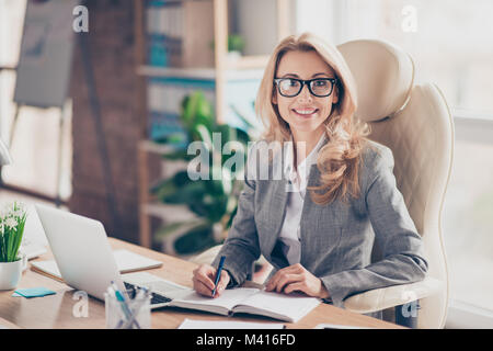 Ritratto di sorridenti allegro rappresentante fiducioso di indossare abiti formali, lei è seduta davanti al computer e la scrittura di informazioni utili Foto Stock