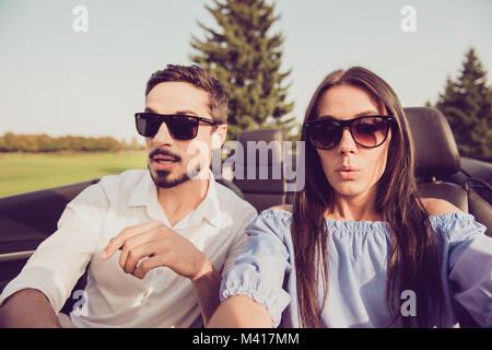 Oh no! Wtf! Close up Gorgeous Brunette lady driver e il suo brunet guy nel panico, entrambi ben vestito in nero gli occhiali trendy, parco con alberi verdi Foto Stock