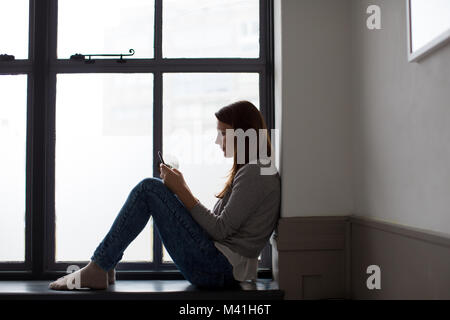 Giovane femmina adulta seduto sul davanzale con lo smartphone Foto Stock