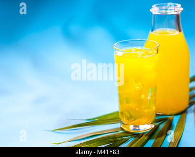 Paradiso tropicale concetto: una bottiglia di luminose e colorate di succo d'arancia con un bicchiere di succo di frutta pieno di cubetti di ghiaccio su un sfondo turchese con un palmare Palm Foto Stock