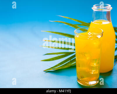 Paradiso tropicale concetto: una bottiglia di luminose e colorate di succo d'arancia con un bicchiere di succo di frutta pieno di cubetti di ghiaccio su un sfondo turchese con un palmare Palm Foto Stock