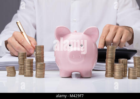 Close-up di imprenditore il calcolo di Bill con Piggybank e monete impilate sulla scrivania in ufficio Foto Stock