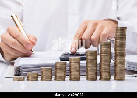 Sezione mediana di imprenditore il calcolo di Bill con la calcolatrice e monete impilate sulla scrivania in ufficio Foto Stock