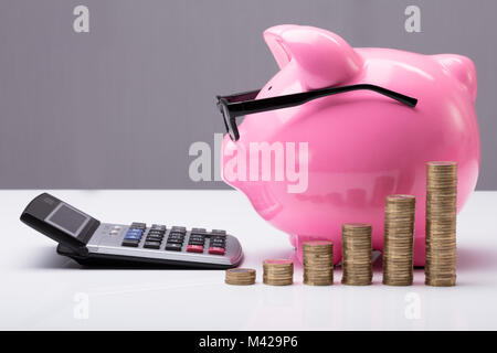 Monete impilate nella parte anteriore del Rosa Salvadanaio con occhiali e Calcolatrice sulla scrivania Foto Stock