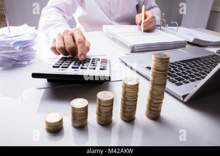 Sezione mediana di un imprenditore calcolo delle imposte utilizzando la calcolatrice nella parte anteriore di una pila di monete Foto Stock