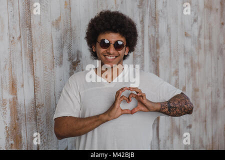 Con l'amore. Positivo e ricci mulatto uomo, indossando occhiali da sole, piegato le mani in una forma di cuore, mentre in piedi contro le vecchie pareti in legno Foto Stock