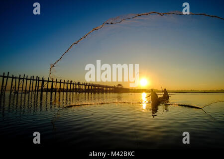 Un pescatore su una barca è di gettare la sua rete nel lago Taungthaman accanto a U Bein Bridge a sunrise Foto Stock