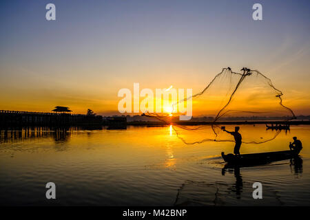 Un pescatore su una barca è di gettare la sua rete nel lago Taungthaman accanto a U Bein Bridge a sunrise Foto Stock