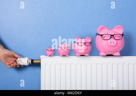 Close-up di mano di una persona di termostato di regolazione con Piggy banche indossando occhiali sul radiatore Foto Stock