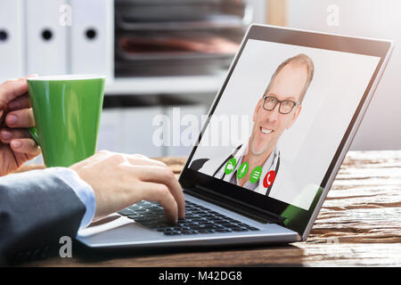 Close-up di mano di una persona le videoconferenze con sorridente medico maschio sulla scrivania in legno Foto Stock