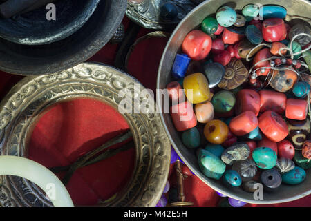 Tibetano gioielli antichi: perle di corallo, turchese e ambra in una ciotola, braccialetti metallici su uno sfondo di colore rosso. Foto Stock