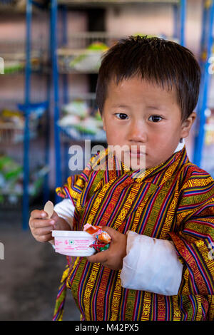 Punakha, Bhutan. Giovane ragazzo in maschio tradizionale indumento Gho, a mangiare il gelato nel mercato Lobeysa. Foto Stock