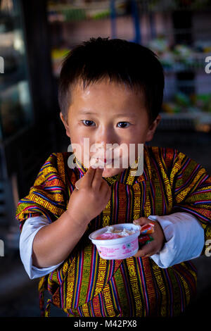 Punakha, Bhutan. Giovane ragazzo in maschio tradizionale indumento Gho, a mangiare il gelato nel mercato Lobeysa. Foto Stock