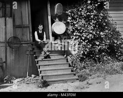 Giovane uomo si siede sulla scalinata anteriore del suo inizio xx secolo americano farm home, ca. 1915. Foto Stock