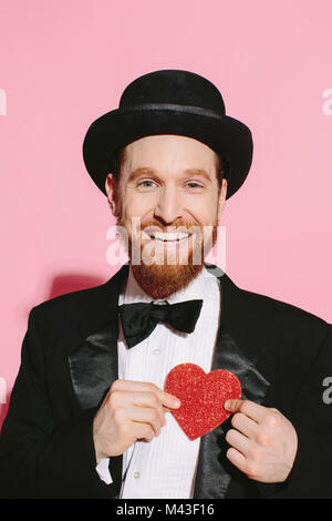 Uomo sorridente in tux e il top hat tenendo un cuore rosso sul suo petto Foto Stock