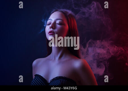 Asian giovane donna di chiudere gli occhi e posa contro uno sfondo di rosso e di blu fumo da un vape su un nero sfondo isolato Foto Stock