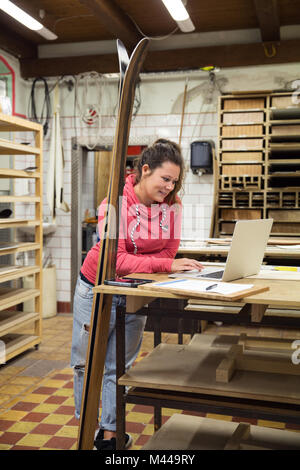 Donna in officina, utilizzando laptop, sci appoggiata contro accanto a lei Foto Stock