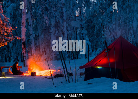 Uomo seduto accanto al fuoco, di notte, nei pressi di tenda in coperta di neve la foresta, Russia Foto Stock