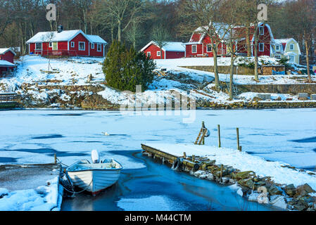 Red case di legno sull'altro lato di una baia congelati. Piccola barca congelato dal jetty in primo piano. Ubicazione Bokevik fuori Ronneby nel sud sw Foto Stock