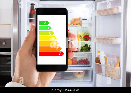 Primo piano della mano d'uomo utilizzando energia etichetta sul telefono cellulare contro il frigorifero a casa Foto Stock